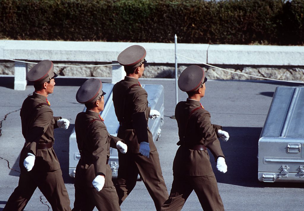 North Korean soldiers in brown uniforms march in formation on an army base complex.