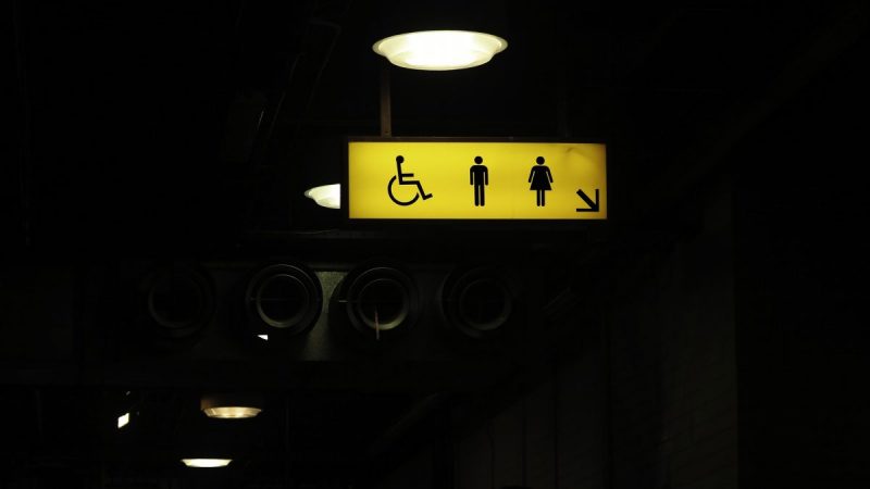 A dim street, with a bright yellow sign hanging above, displaying the symbols for disabled, male, and female toilet facilities.