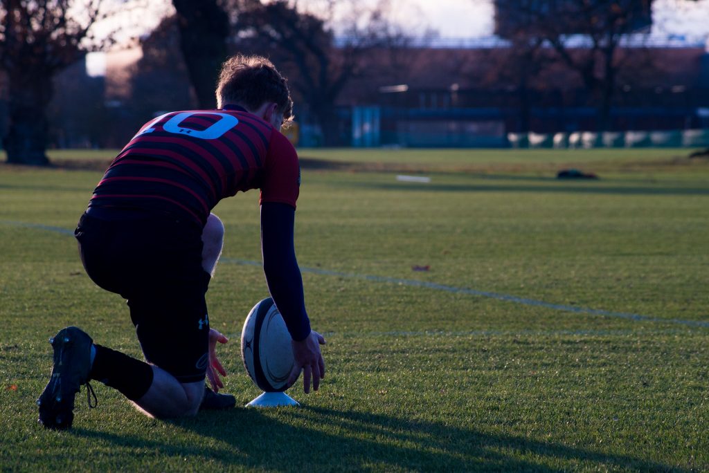 A rugby player kneels on a green pitch, reaching for a rugby ball.