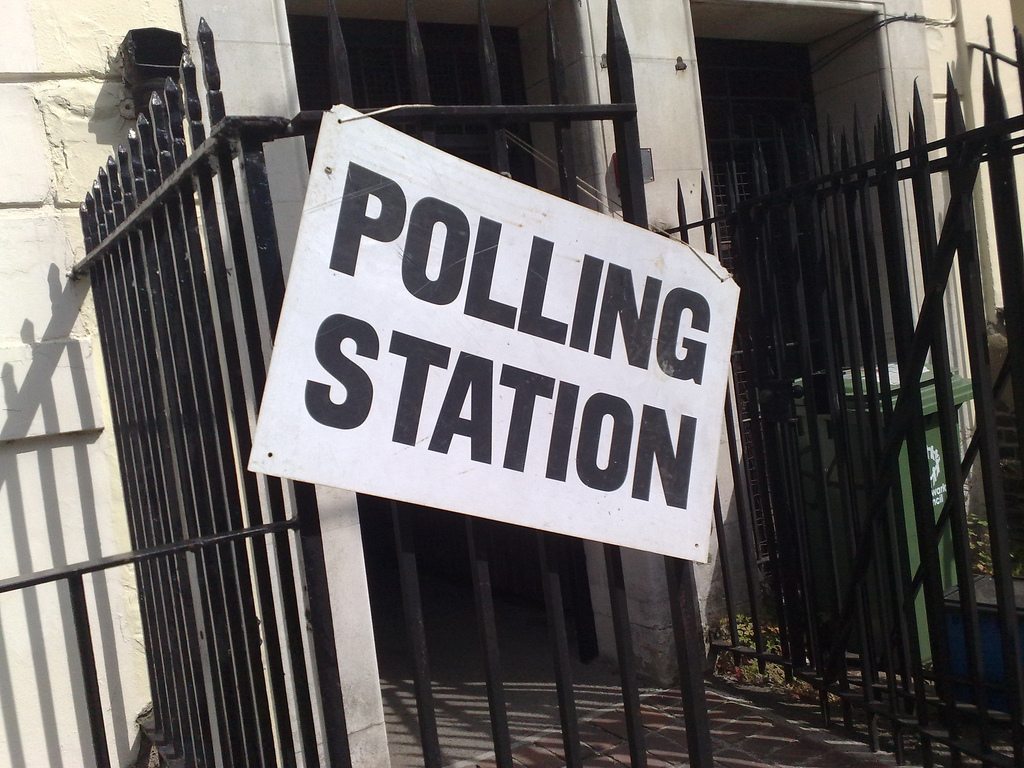 Black railings outside of a white building, with a white "Polling Station" sign hung on the fence.
