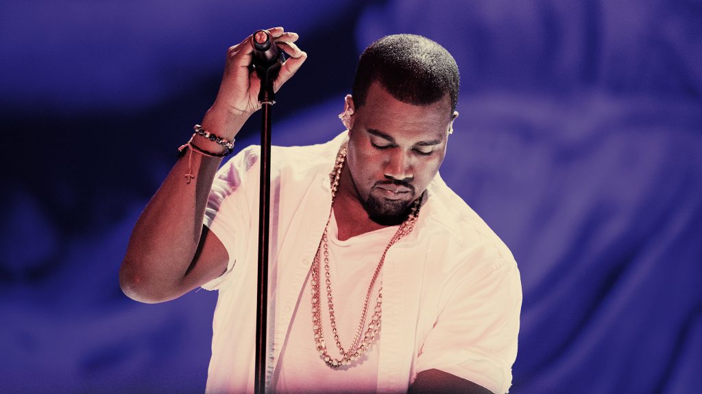 Kanye west wearing a white shirt and gold chains, holding onto a microphone stand with his right hand.