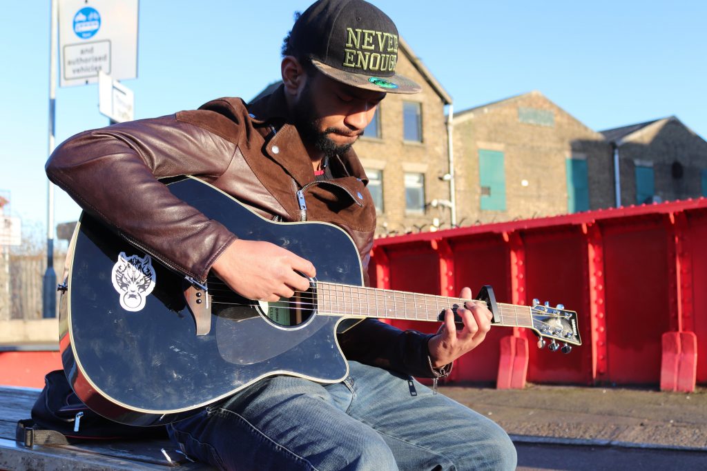 A person with a dark beard, black cap and brown leather jacket plays a black acoustic guitar.